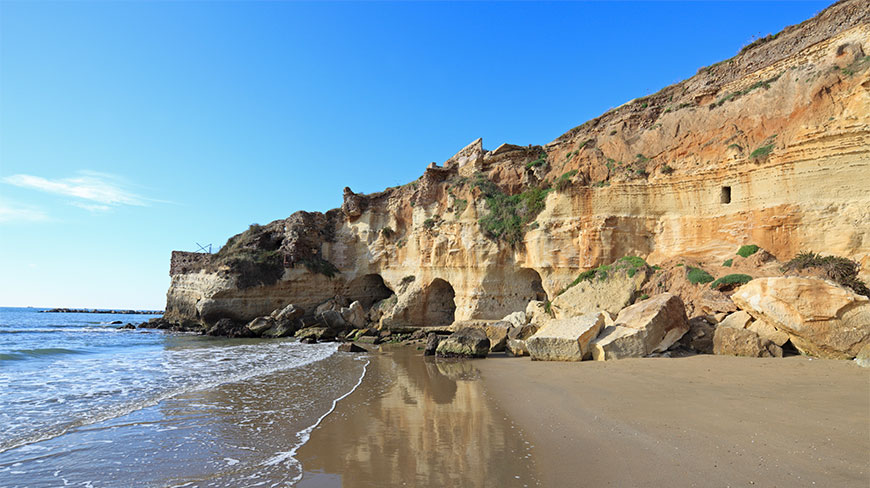 A sandy beach in Anzio, Italy, featuring layered golden cliffs with cave openings under a clear blue sky.
