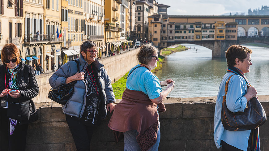 A group of women stands on a bridge in Florence, Italy, with the Arno River and Ponte Vecchio in the background.
