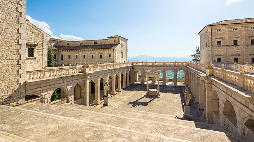 View of the grand stone courtyard at Montecassino Abbey in Cassino, Lazio, featuring arched loggias, statues, and a central well under a clear blue sky.