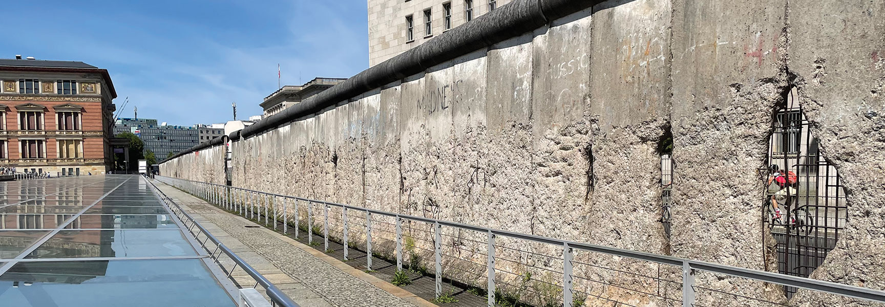 A weathered, preserved segment of the concrete Berlin Wall with holes and damage, standing at an outdoor memorial site under a clear blue sky.
