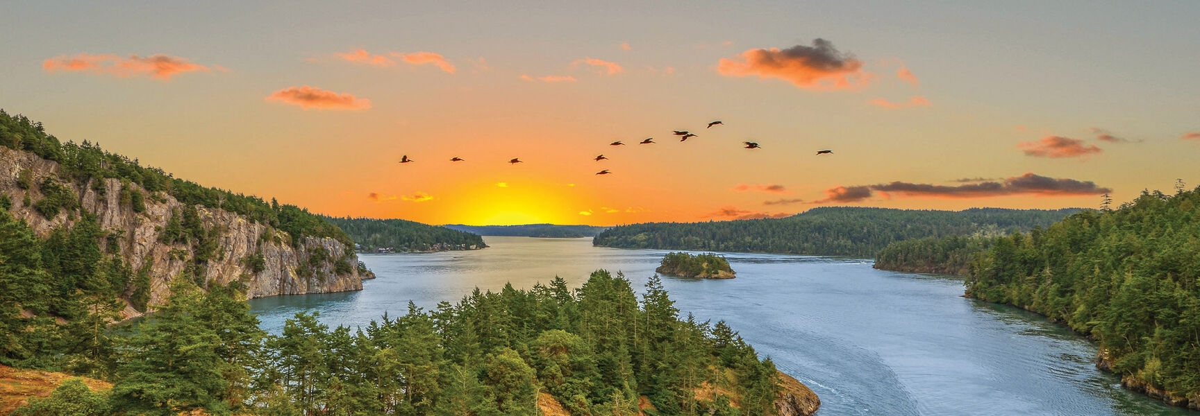 Birds fly over a scenic waterway at sunset, surrounded by the lush evergreen forests and rocky cliffs of Washington's breathtaking Pacific Northwest.