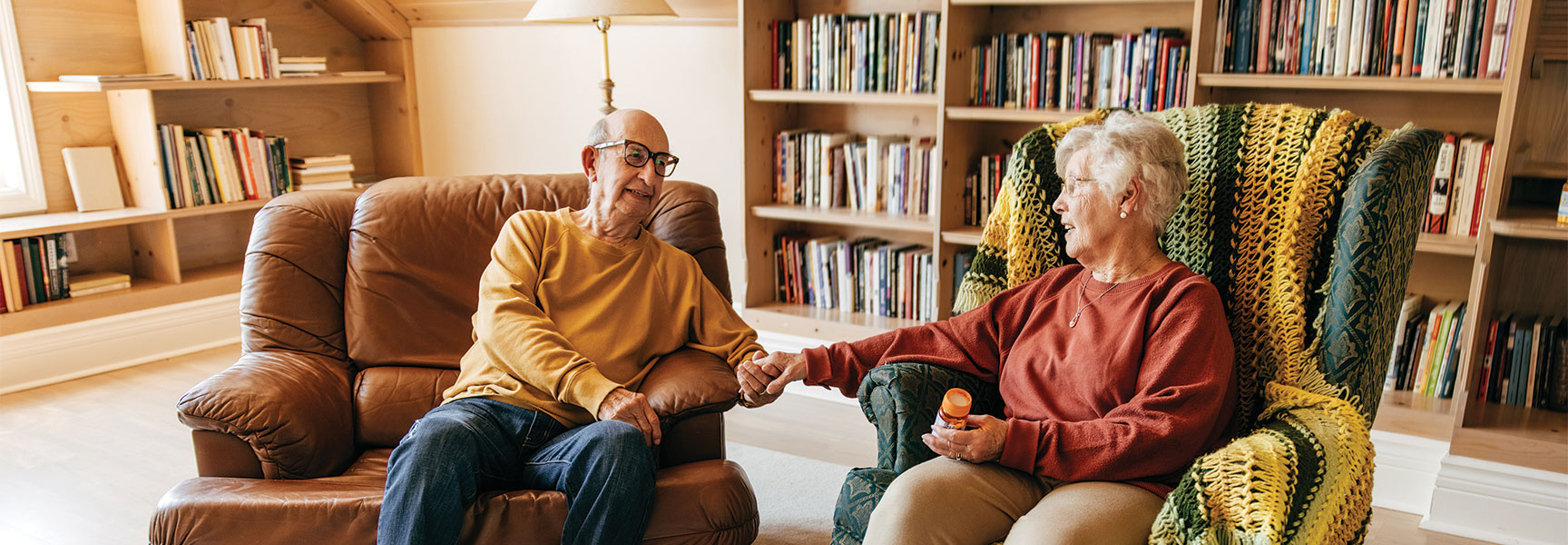 An elderly couple sits in armchairs holding hands in a bookshelf-lined room, illustrating safety and comfort while aging in place.