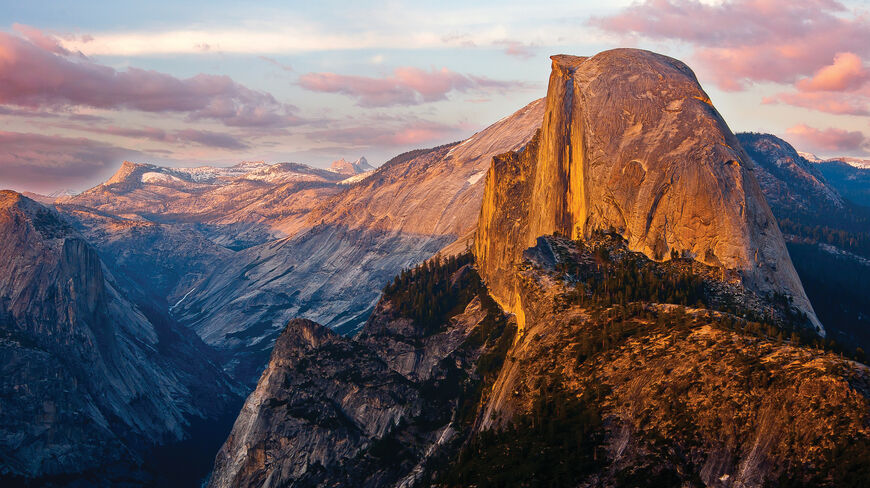 Half Dome in Yosemite National Park, California, glows with golden sunset light against a backdrop of rugged mountains and soft pink clouds.