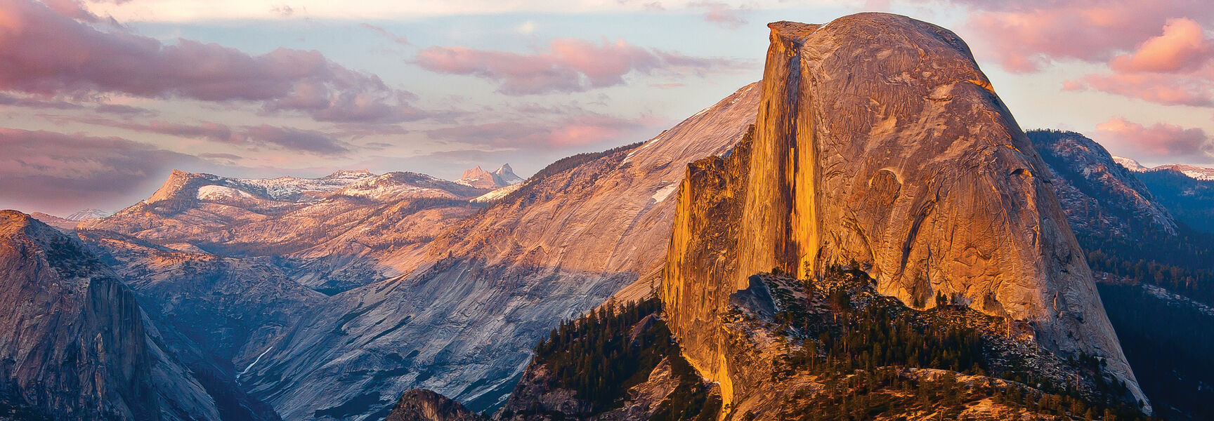 Half Dome in Yosemite National Park, California, glows with golden sunset light against a backdrop of rugged mountains and soft pink clouds.