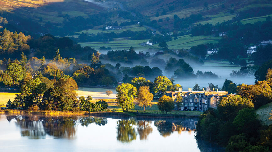 Scenic landscape of England’s Lake District featuring a stone building reflected in a calm, misty lake surrounded by rolling green hills.