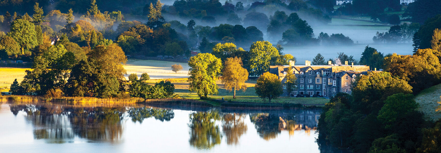 Scenic landscape of England’s Lake District featuring a stone building reflected in a calm, misty lake surrounded by rolling green hills.