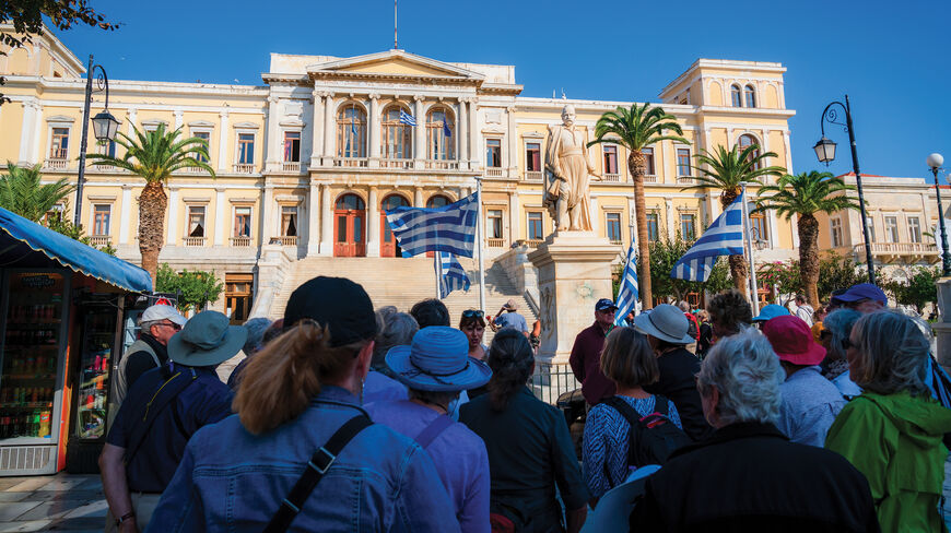 Tourists gather before a grand neoclassical building with Greek flags during a cultural voyage through history in the Greek Isles.