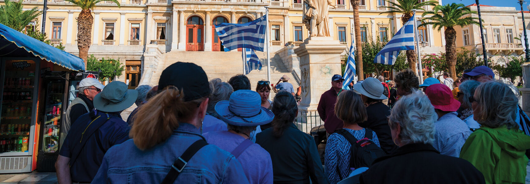 Tourists gather before a grand neoclassical building with Greek flags during a cultural voyage through history in the Greek Isles.