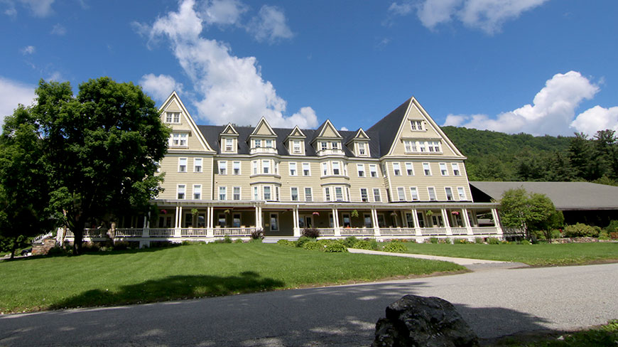The large, tan Silver Bay YMCA building with a long covered porch, viewed from a lawn on a bright, sunny day.