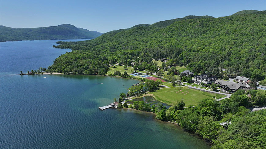An aerial US NY view looking south over a lakeside resort nestled between lush green mountains and clear blue waters under a sunny sky.