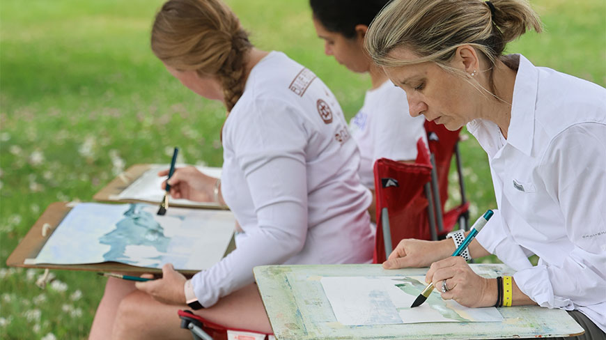Three women sit outdoors in a grassy field in New York, painting landscapes with watercolors on wooden lap desks.