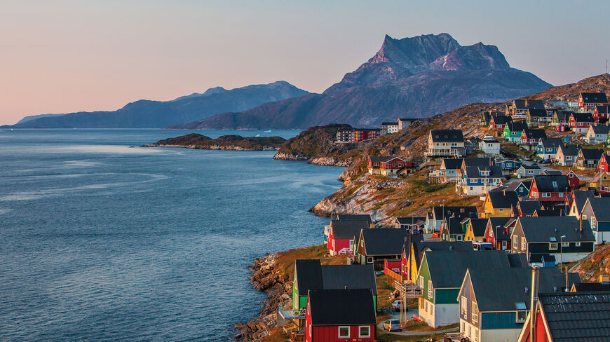 Colorful houses line the rocky coast of a Greenland village, with a large mountain peak rising above the blue water in the background.