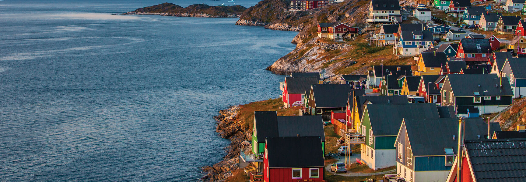 Colorful houses line the rocky coast of a Greenland village, with a large mountain peak rising above the blue water in the background.
