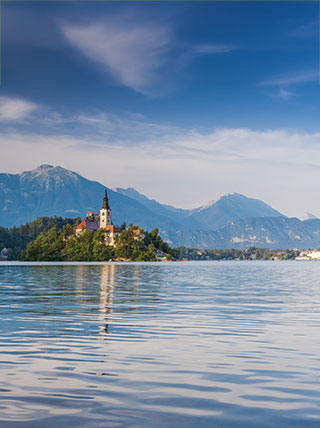 A church on a small island in a lake with a mountain range in the background under a partly cloudy sky.