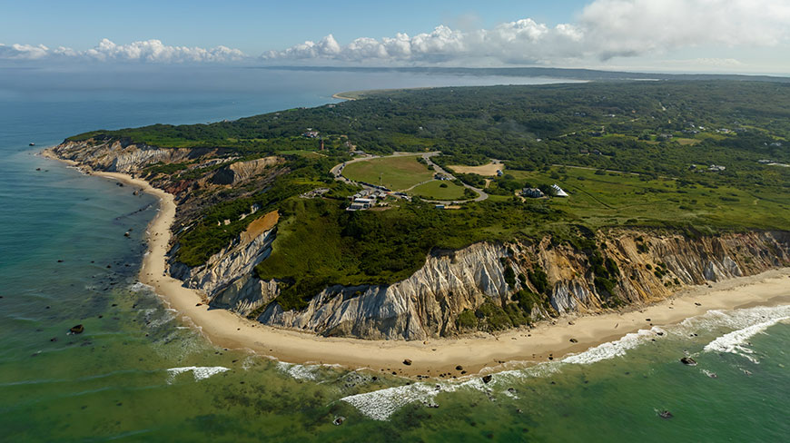 An aerial view of the colorful clay cliffs and lush landscape of Martha's Vineyard, Massachusetts, meeting the calm ocean on a sunny day.