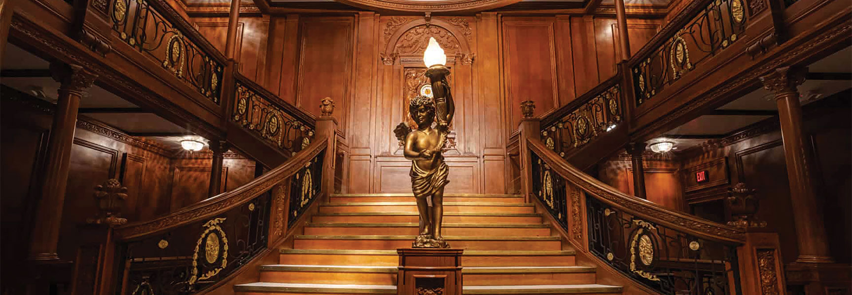 Ornate replica of the Titanic's Grand Staircase with wood paneling and a cherub statue at Titanic: The Artifact Exhibition.