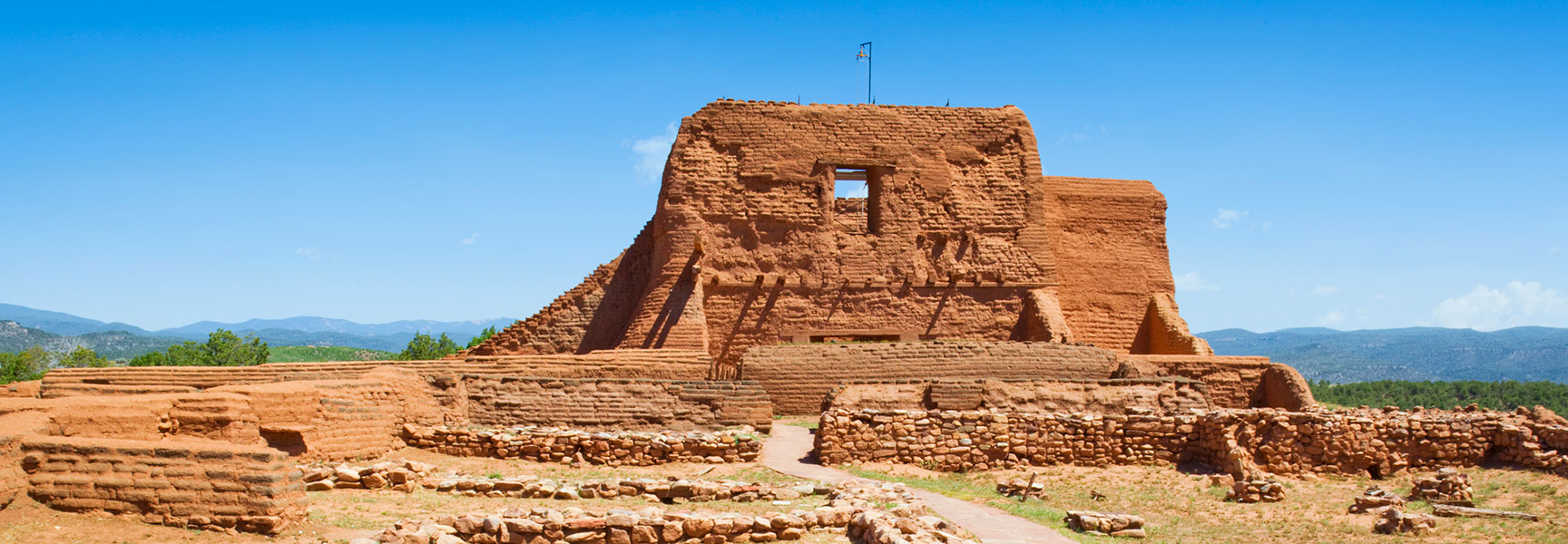 The reddish-brown adobe ruins of the Pecos Mission Church stand under a bright blue sky at Pecos National Historical Park in New Mexico.