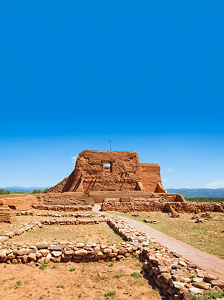 An adobe mission ruin at Pecos National Monument stands in a field of stone foundations beneath a bright blue sky.