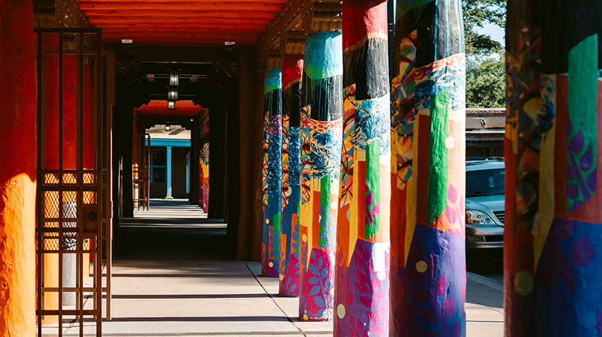 A row of vibrantly painted, multicolored pillars lines a sunlit covered walkway, receding into the distance.