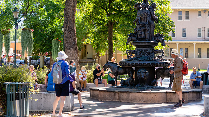 A group of tourists gathers around an ornate bronze water fountain in a sunny park with large green trees.