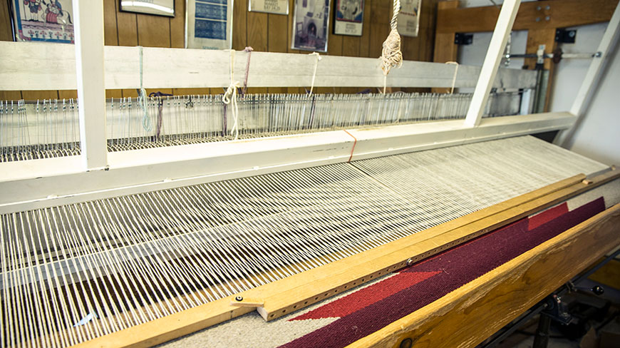 A close-up of a large weaving loom with white threads and a partially finished red textile.