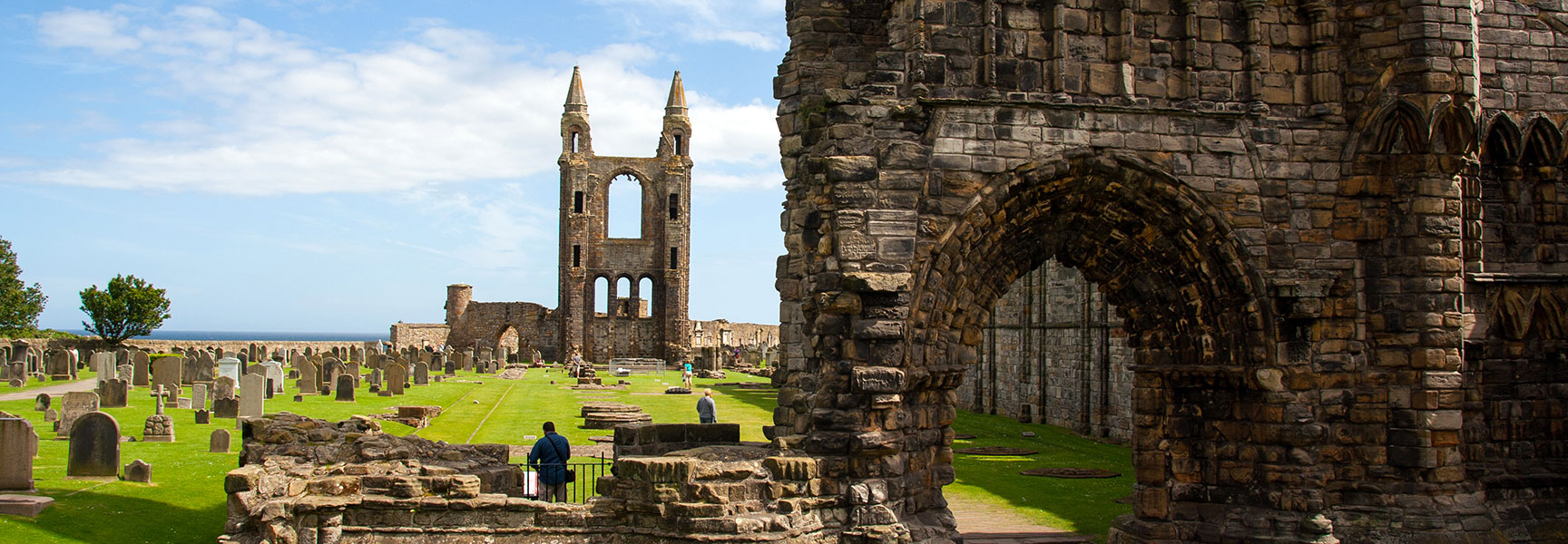 The stone ruins of St Andrew's Cathedral in Scotland, with a graveyard on a green lawn overlooking the sea under a blue, partly cloudy sky.