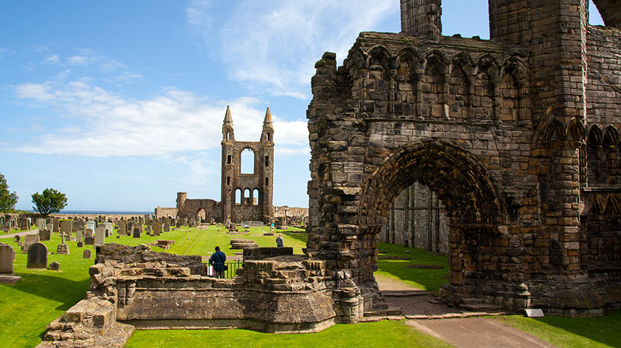 The stone ruins of the Cathedral of St Andrew in Scotland are seen across a green lawn and historic graveyard on a sunny day.