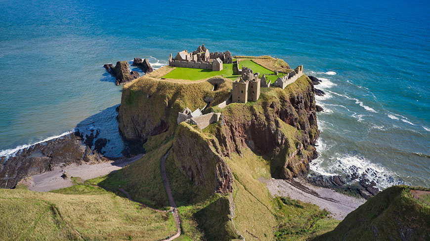The ruins of Dunnottar Castle sit atop a dramatic, grassy cliff overlooking the bright blue sea near Stonehaven, Scotland.