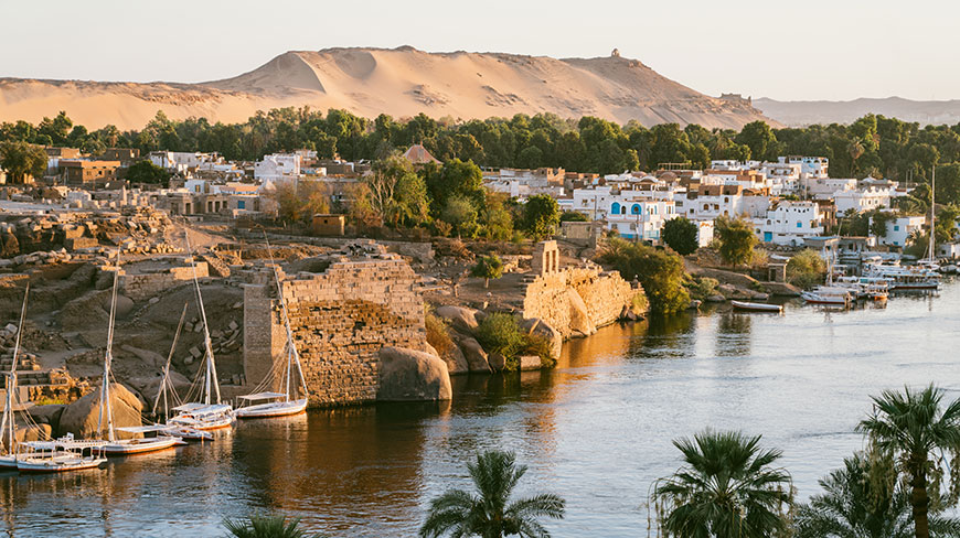 Sailboats docked on the banks of the Nile River in Aswan with ancient ruins and a village in front of a sand dune.