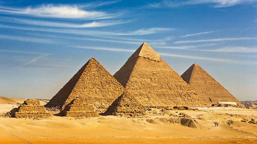 The great pyramids of Giza sit in the sandy Egyptian desert under a bright blue, cloud-streaked sky.
