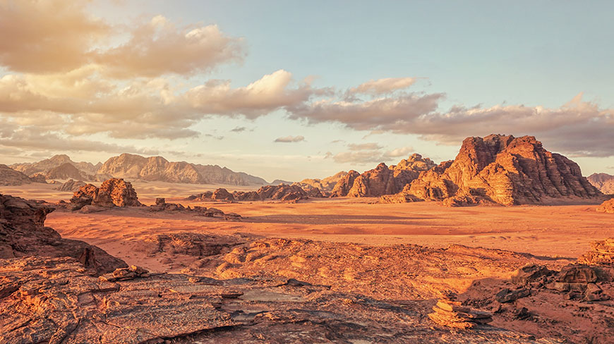 The rocky, red landscape of the Wadi Rum Desert in Jordan stretches out under a partly cloudy, golden-hour sky.