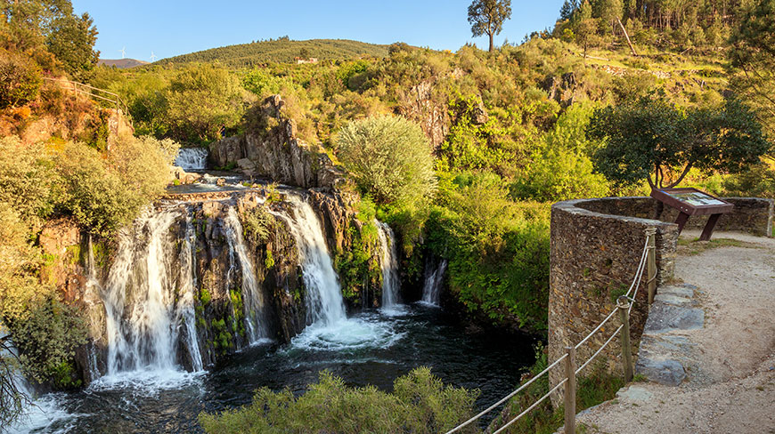 A wide waterfall cascades over rocks into a pool, with a stone viewing area, surrounded by the lush green landscape of Costa Rica.