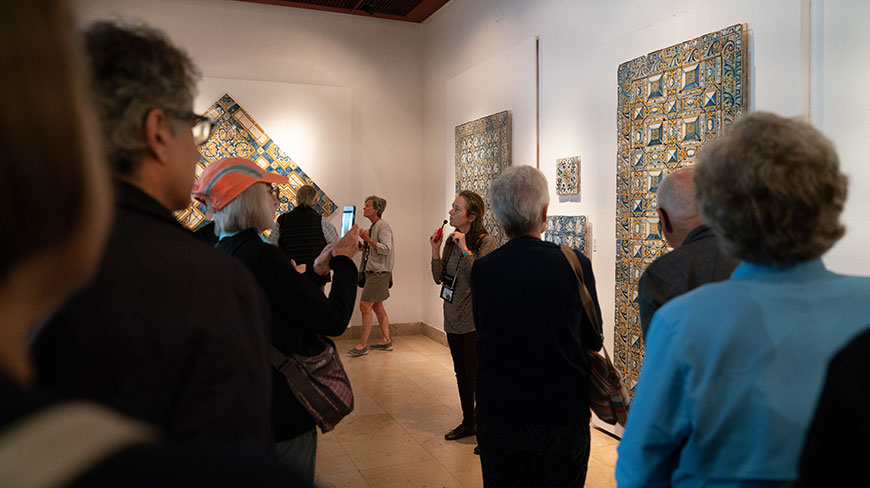 A tour guide speaks to a group of visitors in a museum gallery with decorative blue and yellow tiles displayed on the wall.