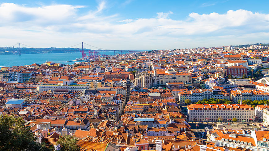An aerial view of the historic Lisbon cityscape with its iconic red roofs, overlooking the Tagus River and 25 de Abril Bridge under a blue sky.