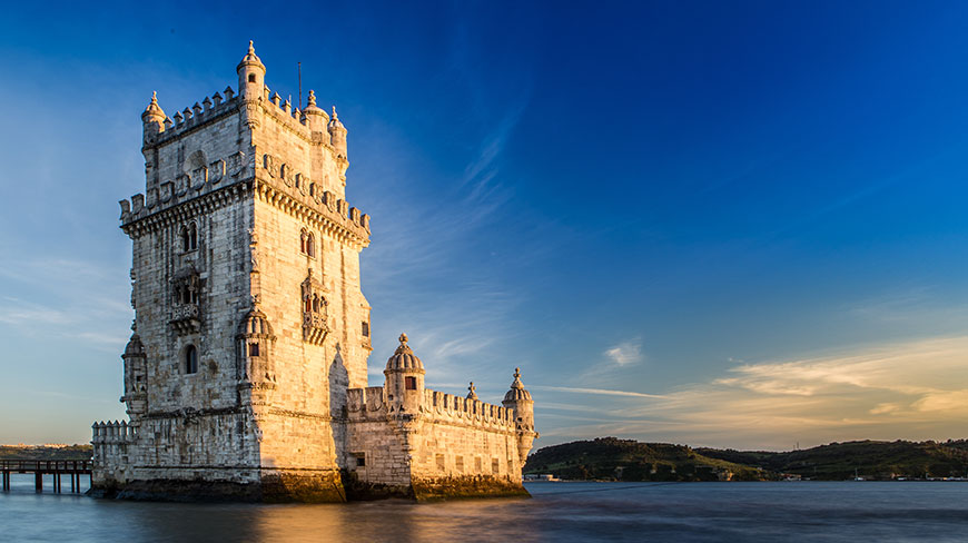 Lisbon's historic Belém Tower sits at the water's edge under a clear blue sky during a beautiful golden hour.