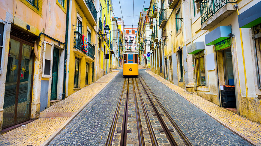 A classic yellow tram travels up a steep, narrow cobblestone street lined with colorful, weathered buildings in Lisbon.