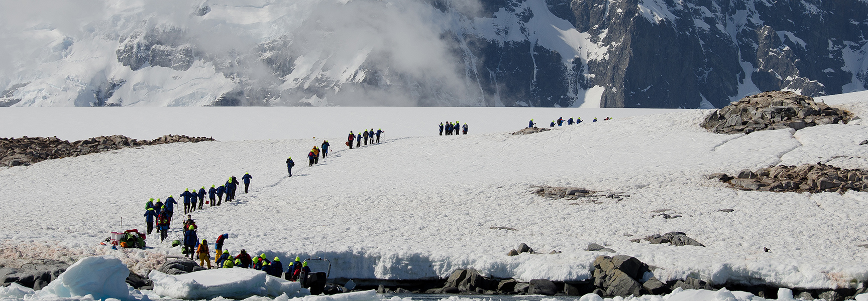 A large group of travelers hike up a snow-covered hill in a long line with dramatic, snow-covered mountains in the background.