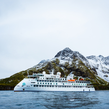 The Greg Mortimer expedition ship floats in the water in front of a large, snow-dusted mountain range under an overcast sky.