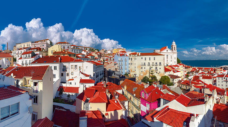 A sunny cityscape of Alfama, Lisbon, with red-tiled roofs, a bright blue sky, and the coast in the distance.