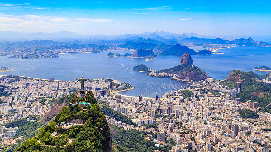 An aerial view of Rio de Janeiro shows the Christ the Redeemer statue overlooking the city, with Sugarloaf Mountain and Guanabara Bay in the distance.