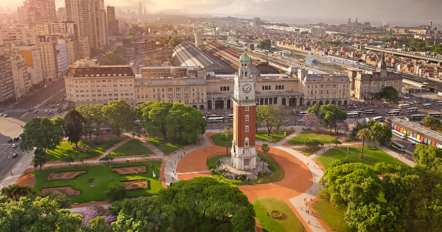 An aerial view of a historic clock tower surrounded by a green park and the Buenos Aires cityscape at sunset.