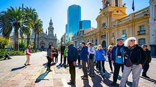A group of travelers listens to a tour guide in a sunny plaza in downtown Santiago, Chile, with historic and modern buildings behind them.