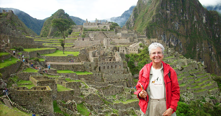 A woman in a red jacket smiles in front of the ancient stone ruins of Machu Picchu nestled in the lush green mountains.