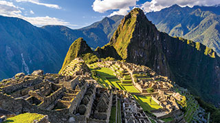 The ancient stone ruins of Machu Picchu sit on a green terrace with a dramatic, steep mountain peak rising in the background.