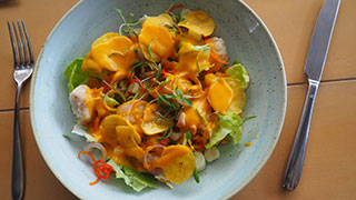 A light blue bowl holds a serving of ceviche with a yellow sauce, garnished with fresh greens, with a fork and knife beside it.