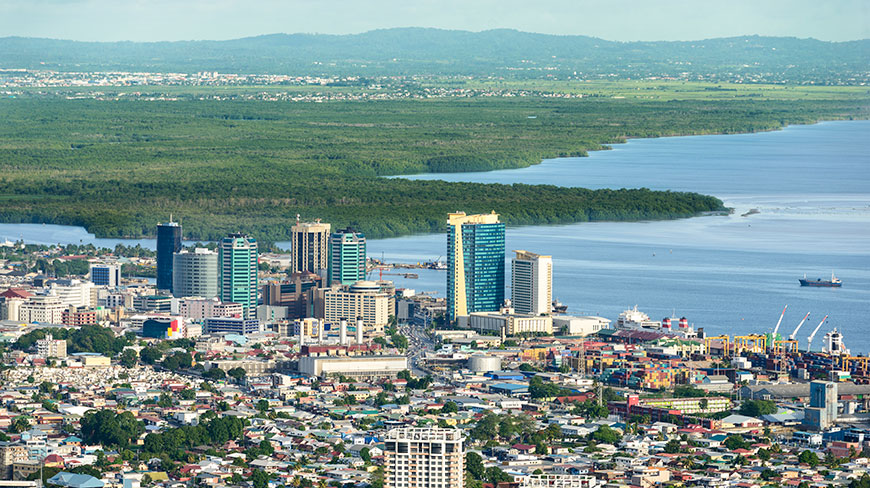 An aerial view of Port of Spain, Trinidad, with its downtown skyline and harbor situated between the blue ocean and a lush green landscape.