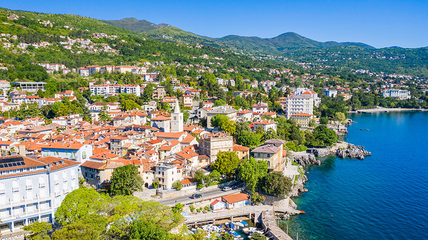 An aerial view of the coastal town of Lovran in Croatia, with its red-roofed buildings lining the deep blue Kvarner Bay.