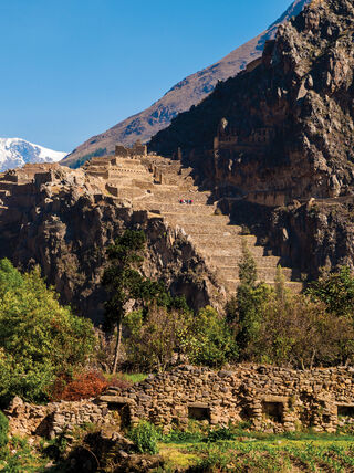 Ancient stone terraces and ruins in the Sacred Valley of Peru, with steep mountains and a snow-capped peak under a clear blue sky.