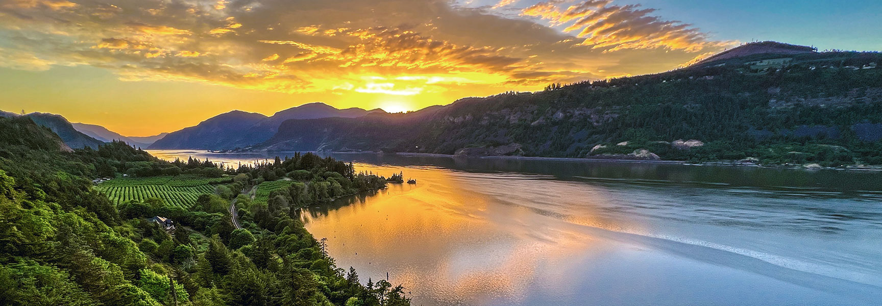 A vibrant sunset over the Columbia River Gorge in Oregon, with golden light reflecting on the water and lush green hillsides.