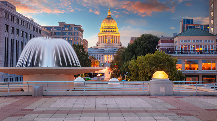 View of the illuminated Wisconsin State Capitol dome at sunset from a plaza with a large white fountain in Madison.
